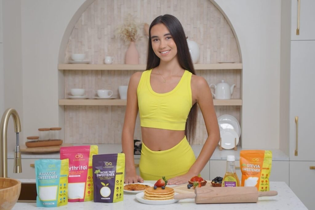 Young woman in a yellow activewear set standing in a modern kitchen, displaying a range of Livsmart natural sweeteners including Monk Fruit, Erythritol, Allulose, and Stevia