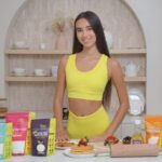 Young woman in a yellow activewear set standing in a modern kitchen, displaying a range of Livsmart natural sweeteners including Monk Fruit, Erythritol, Allulose, and Stevia