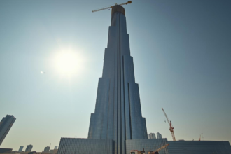 A low-angle shot of the Jeddah Tower under construction in early 2026, showing active cranes on the 80th floor and the glass facade rising against the blue sky